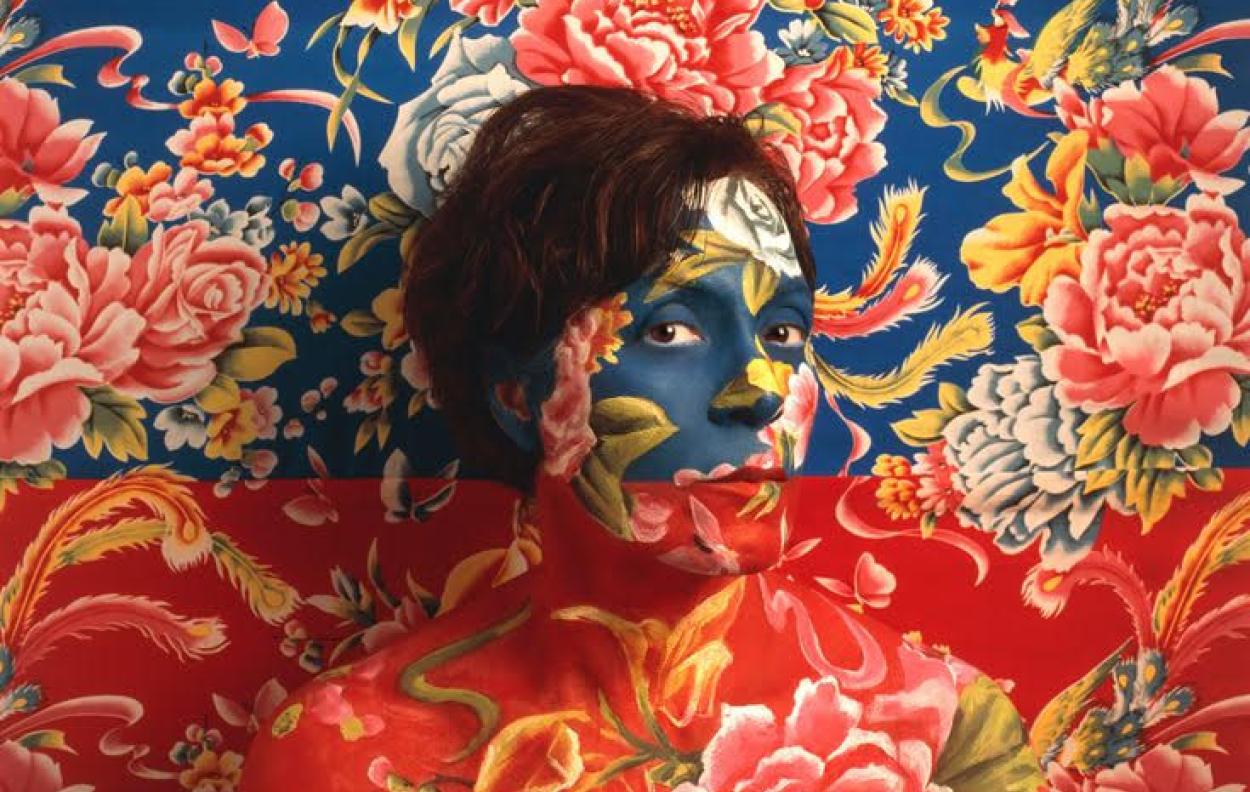 Self Portrait of Artist wearing blue and red floral make-up as camouflage with wallpaper behind her